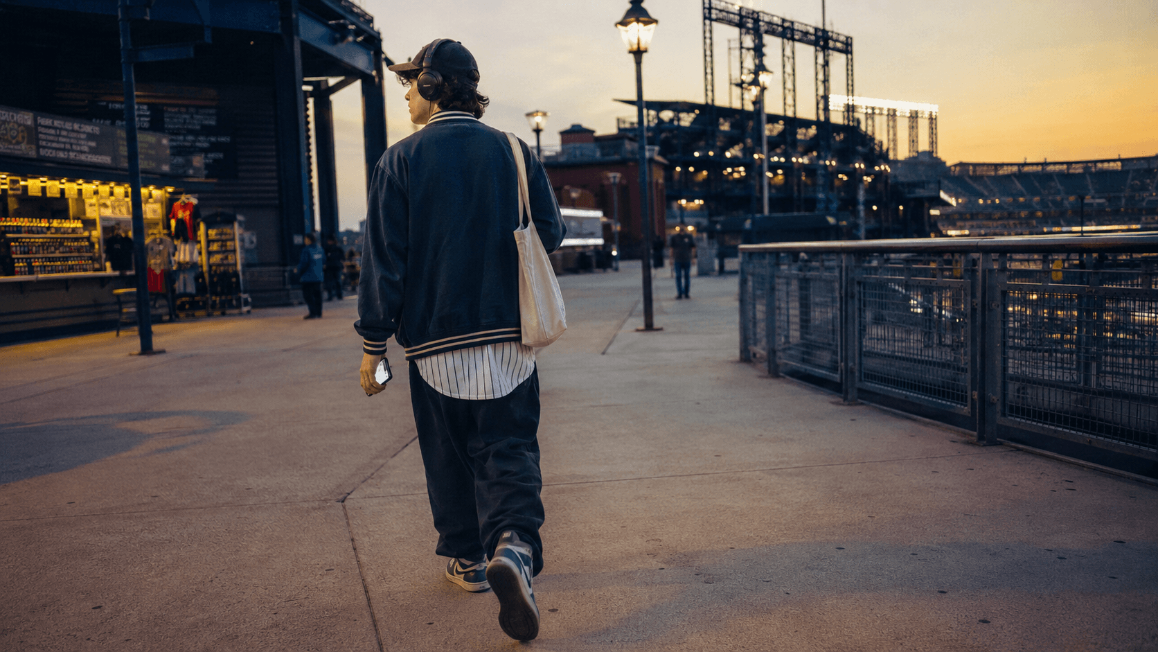 A person walking toward a ballpark, wearing headphones.